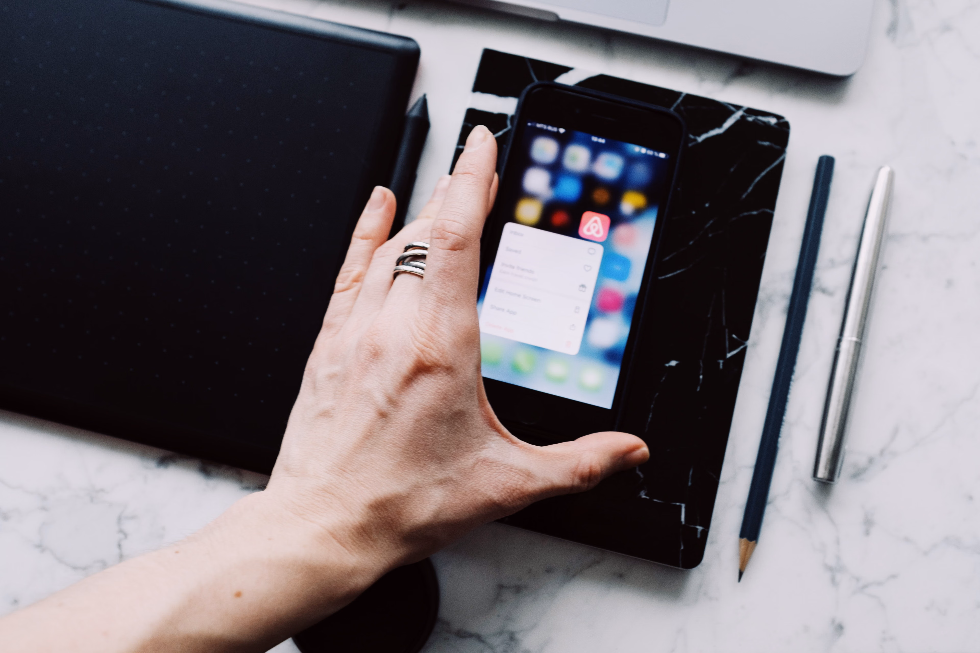Hand holding a smartphone displaying apps on a marble desk with a pen and pencil nearby