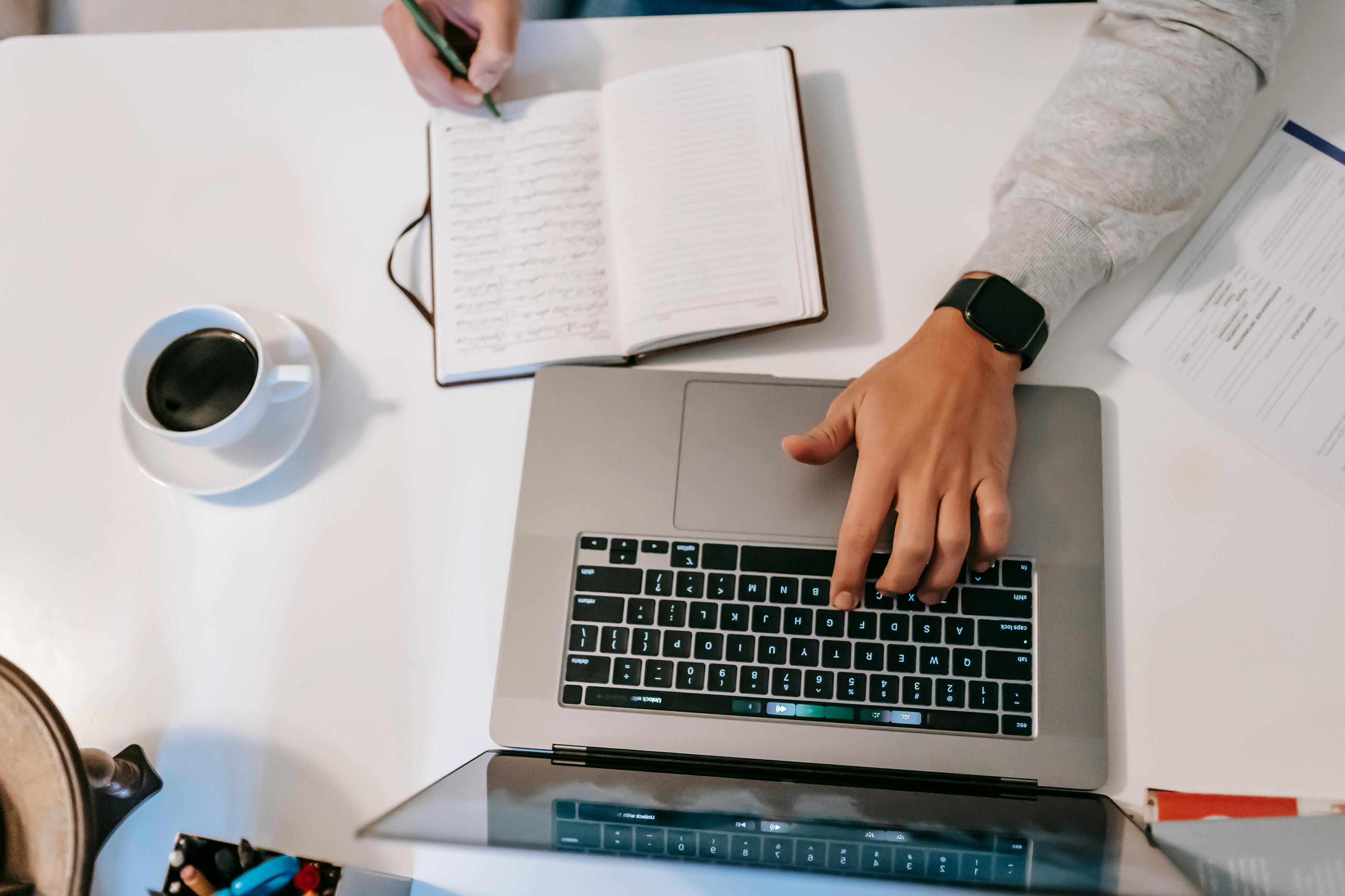 Freelancer typing on laptop with journal and coffee on desk from above